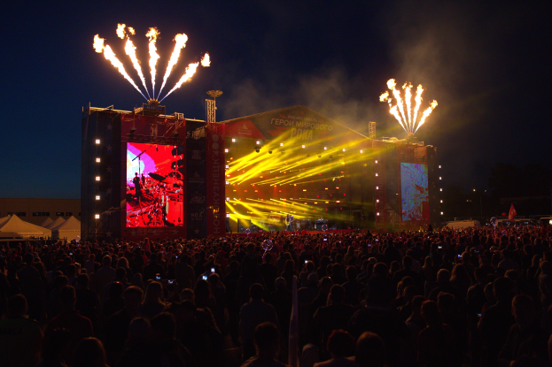 Eine große Menschenmenge versammelte sich auf einem Musikfestival mit einer Bühne in der Mitte, auf der Musiker Instrumente spielten, beleuchtet von hellen Lichtern und umgeben von Bannern, Zelten links und einem dunklen Himmel mit Feuerwerk im Hintergrund.
