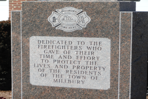 Denkmalstein mit einer Gedenktafel für Feuerwehrleute in Millbury, vor einem Backsteingebäude mit umgebender Vegetation gesetzt.