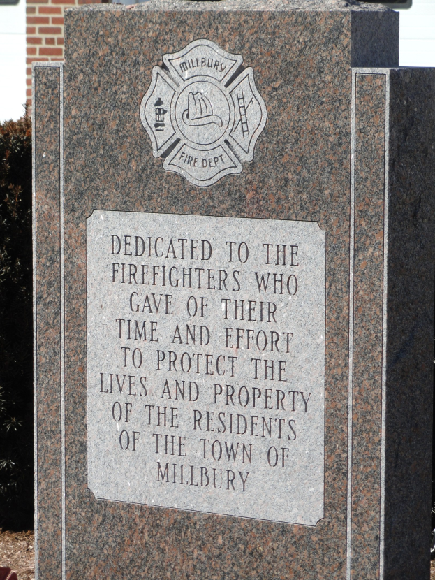 Denkmalstein mit einer Gedenktafel für Feuerwehrleute in Millbury, vor einem Backsteingebäude mit umgebender Vegetation gesetzt.