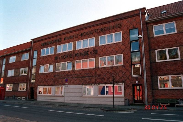 Ein großes Backsteingebäude mit vielen Fenstern, das als Hauptquartier der deutschen Bundespolizei identifiziert wurde, das sich auf einer Straße mit einem Gehweg, einem Baum, einer Bank, einem Laternenpfahl und einem roten Kasten befindet, mit dem Himmel im Hintergrund.