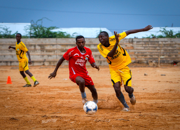 Eine Gruppe von Männern in bunten Trikots, die auf einem sonnendurchfluteten Fußballfeld spielen, umgeben von einer Wand, Bäumen und einem klaren blauen Himmel.