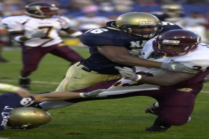 Zwei Fußballspieler in Helmen und Uniformen kollidieren auf dem Feld, während andere im Hintergrund beobachten.