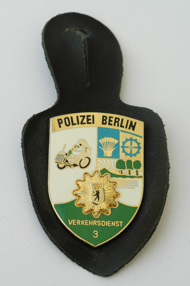 Ein schildförmiges Polizeisiegel mit "Polizei Berlin" in schwarzer fetter Schrift auf einem blauen Hintergrund mit einem weißen Stern und Rahmen, das auf einer Oberfläche ruht.