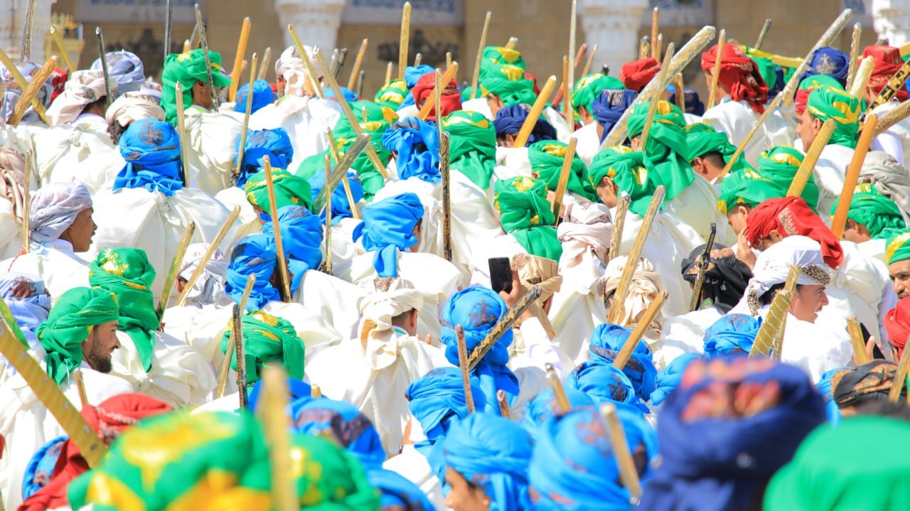 Eine große Gruppe von Menschen in farbenfrohen Kostümen, die eine Straße entlangmarschieren und jeweils einen Stock halten, während sie ein Gebäude im Hintergrund haben.