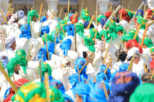 Eine große Gruppe von Menschen in farbenfrohen Kostümen, die eine Straße entlangmarschieren und jeweils einen Stock halten, während sie ein Gebäude im Hintergrund haben.
