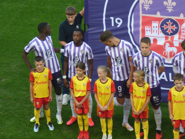 Gruppe junger Menschen auf einem saftig grünen Fußballfeld mit einem "Ligue 1"-Banner im Hintergrund, einige tragen Trikots und andere Shorts.