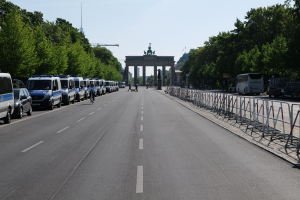 Eine Reihe von Polizeiwagen auf einer Straße vor dem Brandenburger Tor in Berlin, Deutschland geparkt, mit Menschen auf Fahrrädern und Stehenden, Barrieren, Bäumen und einem Bogen mit Statuen im Hintergrund.