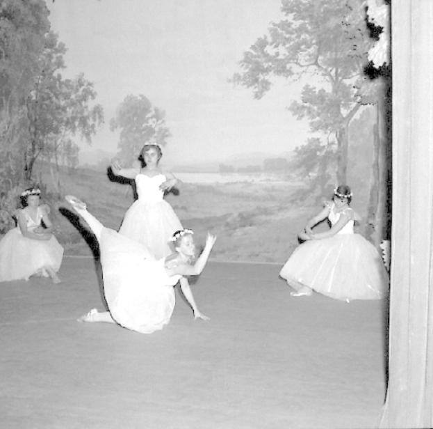 Vier junge Balletttänzerinnen in weißen Kleidern tanzen auf einer Bühne mit einer gemalten Kulisse aus Bäumen und Himmel, dargestellt in Schwarz-Weiß.