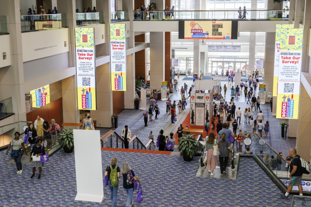 Eine große Menschenmenge bewegt sich durch ein großes Gebäude mit Bannern, Topfpflanzen, Rolltreppen, Geländern, Säulen, bedruckten Schildern, Glastüren und anderen Merkmalen, wobei einige Taschen tragen, was auf die Teilnahme an der Nationalen Buchmesse hinweist.