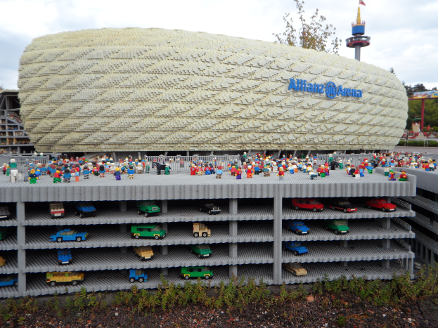 Eine große Lego-Brick-Struktur, die dem Allianz Arena in München, Deutschland, ähnelt, mit einer Gruppe von Menschen davor, umgeben von Bäumen, Pfählen, einem Turm und Grünzeug unten, unter einem bewölkten Himmel.
