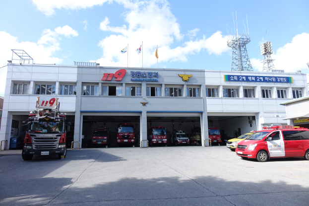 Feuerwache mit mehreren Feuerwehrautos vor dem Gebäude geparkt, Text auf dem Gebäude, Flaggen mit Stangen im Hintergrund, bewölkter Himmel und eine ferne Turm.