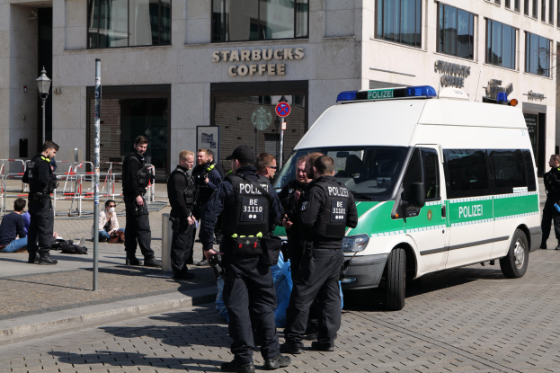 Eine Gruppe von Polizisten steht vor einem Starbucks-Café, rechts davon ein Van und links ein paar Menschen; im Hintergrund sind ein Gebäude, ein Schild, ein Laternenpfahl und ein Zaun zu sehen.