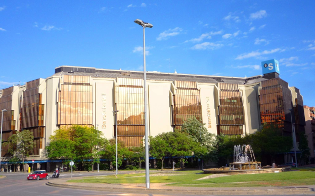 Großes Gebäude namens Bancroft Stadium mit einem Brunnen davor, umgeben von Straßenmasten, Lichtern, Fahrzeugen, Passanten, Bäumen und einem bewölkten Himmel.
