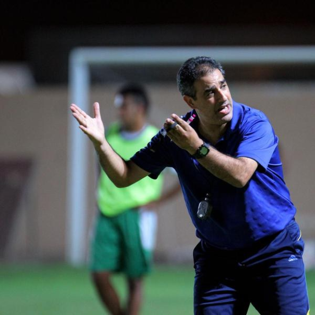 Ein Mann in einem blauen T-Shirt und Hose steht auf einem Fußballfeld und hält ein Mikrofon in der Hand, während er Spielern Zeichen gibt. Im Hintergrund sind eine Person in einem grünen Kleid, ein Torpfosten und eine Wand zu sehen.
