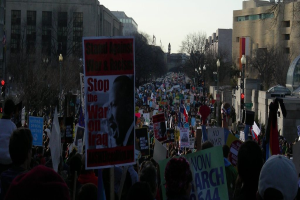 Eine Gruppe von Menschen, die auf einer Straße protestieren und Plakate halten, mit Gebäuden, Bäumen und Laternen im Hintergrund unter einem klaren Himmel.