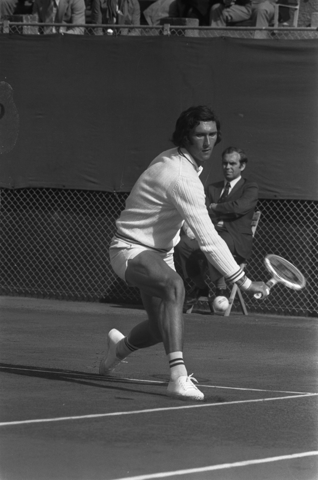 Ein professioneller Tennisspieler in Schuhen schwingt einen Schläger gegen einen Ball während eines Matches, mit Maschendrahtzaun und Zuschauern im Hintergrund.