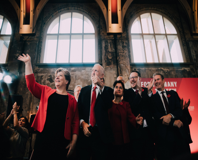 Eine Gruppe von Labour-Parteimitgliedern, die bei einem Wahlsiegereignis jubeln, mit einem Banner und beleuchteten Fenstern im Hintergrund.