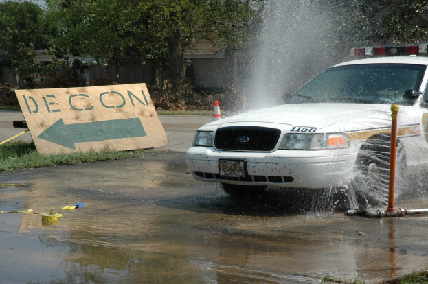 Ein Polizeiwagen fährt durch eine Pfütze, mit einem Schild, einer Verkehrsinsel und anderen Gegenständen am Boden, mit Bäumen, Gebäuden und einem klaren blauen Himmel im Hintergrund.