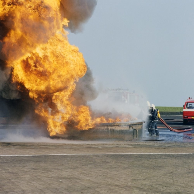 Feuerwehrlöschfahrzeug in Flammen auf der Straßenseite mit zwei Helmträgern, die Stäbe halten, einem Fahrzeug im Hintergrund und dem Himmel.