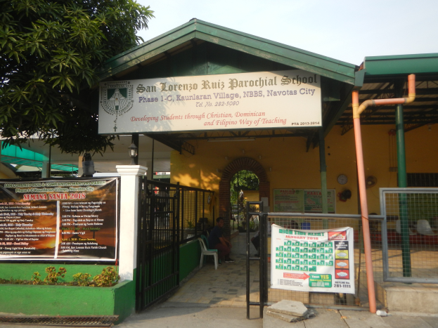 Der Eingang der San Lorenzo Ruiz Pfarrschule zeigt eine Tafel mit Text, einen Metallzaun, Menschen auf Stühlen unter einem Dach, Pfosten, ein Schild, Pflanzen, einen Baum und einen bewölkten Himmel.