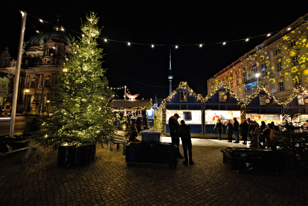 Ein grosser, beleuchteter Weihnachtsbaum steht in der Mitte eines belebten Berliner Marktplatzes, umgeben von Menschen, mit festlichen Dekorationen und Gebäuden im Hintergrund unter einem dunklen Himmel.