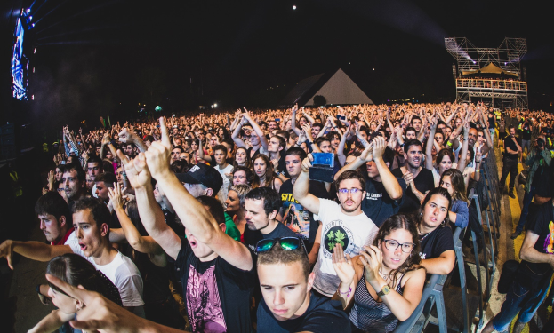 Menschenmenge auf einem Musikfestival mit erhobenen Händen, einige sitzen und andere stehen, neben einem Metallzaun mit Gebäuden und Lichtern im Hintergrund.