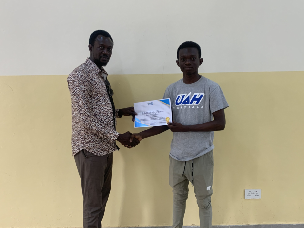 Zwei Männer, die vor einer gelben Wand Hände schütteln, wobei der Mann links ein Zertifikat hält, nachdem er ein Stipendium von der Universität Ghana erhalten hat.