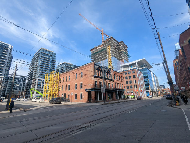 Eine belebte Straße in Toronto mit fahrenden Autos, Passanten auf dem Gehweg, Überlandleitungen auf Masten, Schildern, Gebäuden und einem bewölkten Himmel, mit einem Baugerüst mit einem Kran in der Ferne.