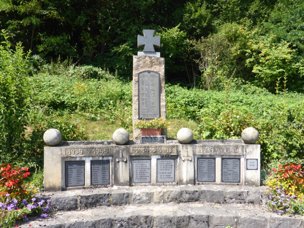 Ein Gedenkstein mit einem Kreuz darauf, beschriftet mit Wörtern und Zahlen, steht in einem Garten umgeben von Pflanzen und Bäumen, wahrscheinlich zum Gedenken an Kriegsopfer.