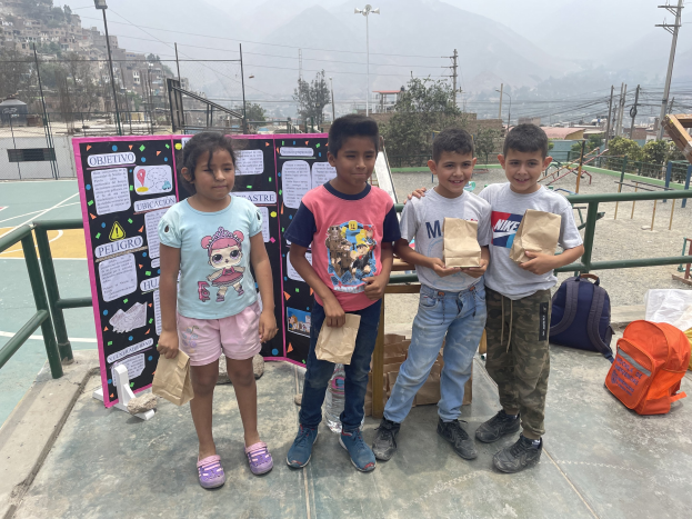 Gruppe von Kindern auf einem Basketballfeld mit Papierbeuteln in den Händen, mit Beuteln und Gegenständen rechts und einer Tafel mit Text links, vor dem Hintergrund von Zäunen, Pfählen, Laternen, Strommasten, Bäumen, Gebäuden, Hügeln und Himmel.