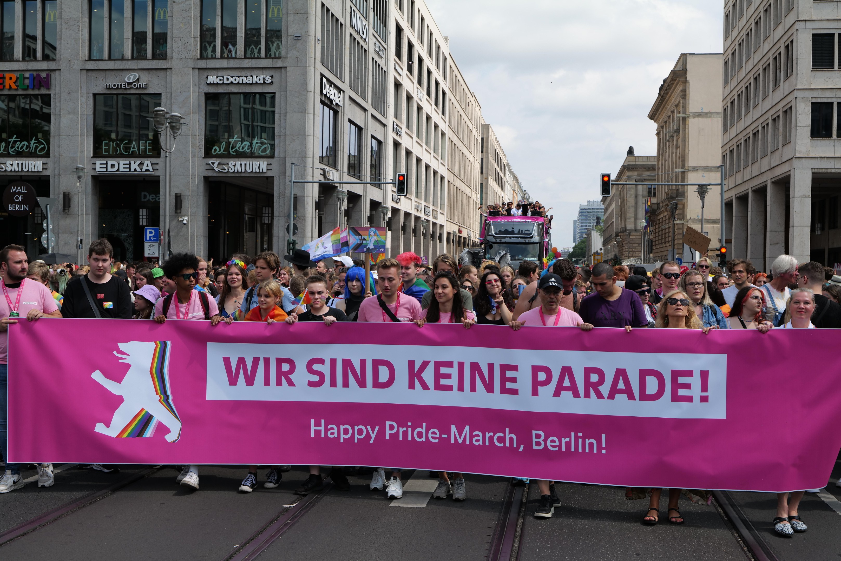 Eine Gruppe von Menschen marschiert auf einer Berliner Straße mit einem pinken Banner, auf dem "Happy Pride March" steht, mit Gebäuden, Laternenmasten und Verkehrszeichen an der Straße unter einem bewölkten Himmel.