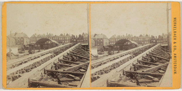 Ein altes Schwarz-Weiß-Foto einer Bahnschiene mit Gebäuden im Hintergrund, das den Text "Merkelbach & Co. Amsterdam" auf der rechten Seite und verstreute hölzerne Gegenstände im Vordergrund zeigt.