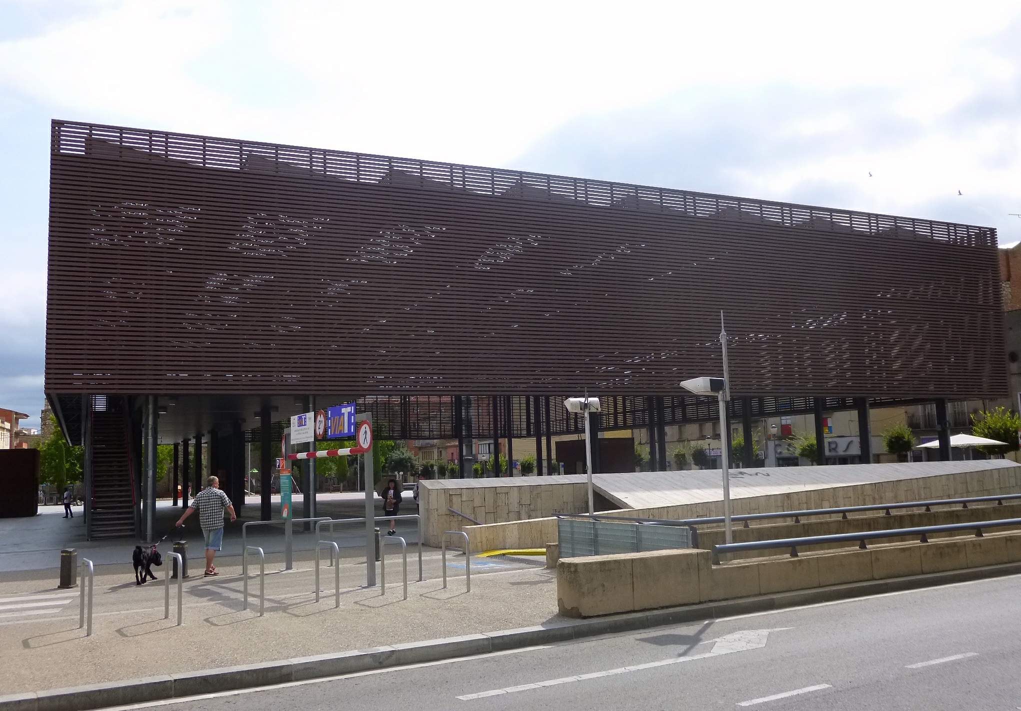 Großes modernes Gebäude mit zahlreichen Fenstern, das als neuer Eingang zum Stadtzentrum dient, umgeben von Straßeninfrastruktur, Fahrzeugen, Fußgängern, Bäumen und einem bewölkten Himmel.