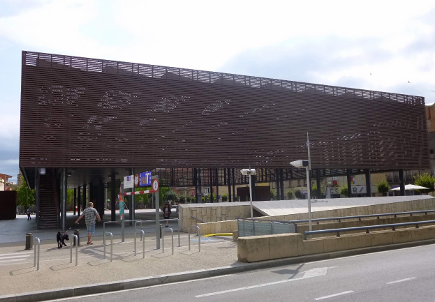 Großes modernes Gebäude mit zahlreichen Fenstern, das als neuer Eingang zum Stadtzentrum dient, umgeben von Straßeninfrastruktur, Fahrzeugen, Fußgängern, Bäumen und einem bewölkten Himmel.