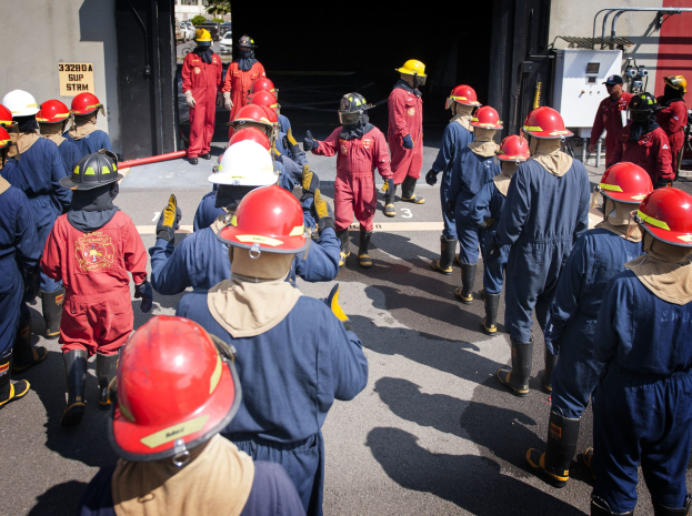 Feuerwehrleute in roter Uniform und Helm, die eine Straße entlanggehen mit einer Wand und Bäumen im Hintergrund unter einem klaren blauen Himmel.