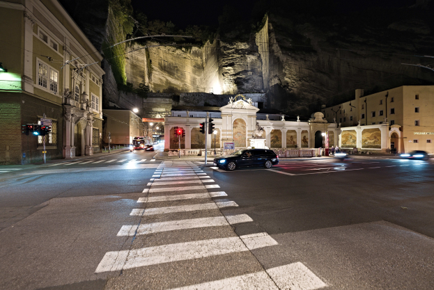 Eine nächtliche Stadtstraße mit fahrenden Autos, Straßenlaternen, Ampeln, Gebäuden mit beleuchteten Fenstern, Laternenpfählen, einem Hügel im Hintergrund und einer Zebrastreifenmarkierung unten.