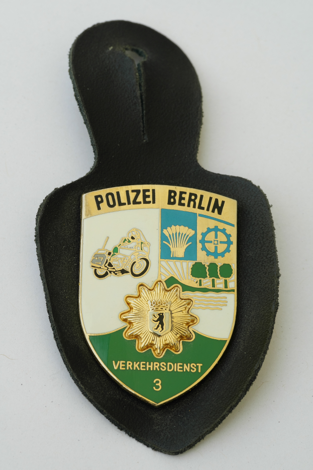 Ein Polizeischild mit der Aufschrift "Polizei Berlin", das ein blaues Schild mit einem weißen Stern in der Mitte zeigt, umgeben von einem weißen Rand, auf einer Oberfläche platziert.