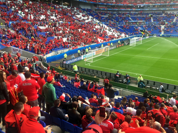 Eine große Menschenmenge in einem Stadion schaut einem Fußballspiel zu, mit Menschen auf Stühlen und Stehplätzen, Fahnen, einem Tor und Deckenbeleuchtung.