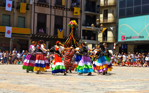 Eine Gruppe Frauen in bunten Kleidern tanzt auf der Straße, umgeben von einer Menge Menschen, mit Gebäuden, Geländern, Balkonen, Fahnen, einem Lichtmast und einer Tafel mit Aufschrift im Hintergrund.
