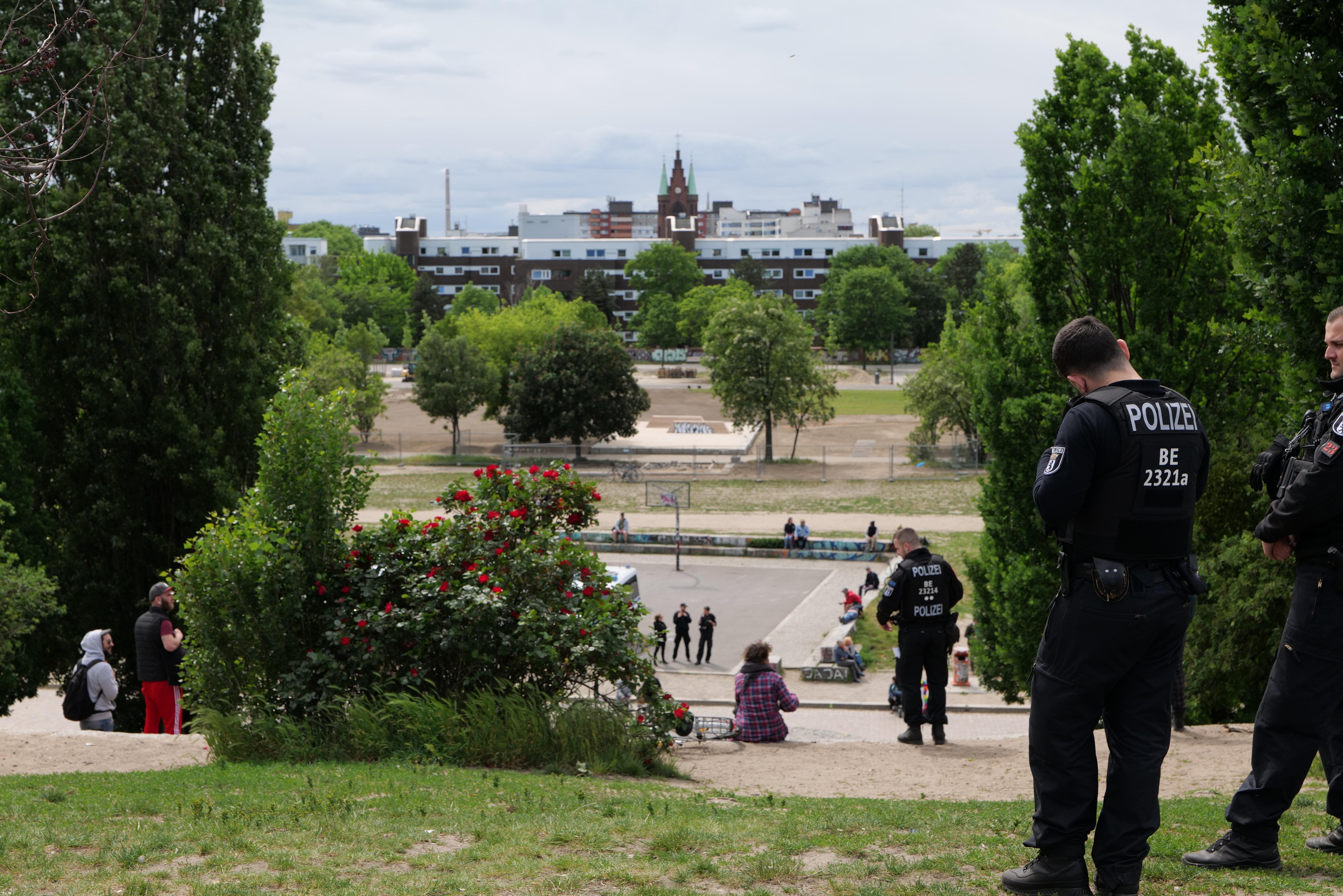 Zwei Polizisten vor einer Gruppe von Menschen in einem Park mit saftigem Grün, Bäumen, bunten Blumen, Gebäuden, Pfählen und einem klaren blauen Himmel.