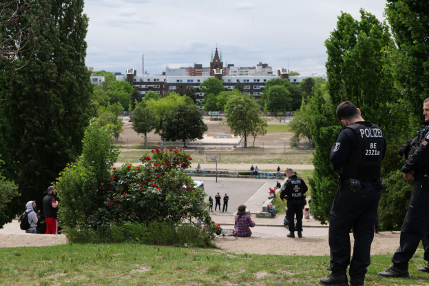 Zwei Polizisten vor einer Gruppe von Menschen in einem Park mit saftigem Grün, Bäumen, bunten Blumen, Gebäuden, Pfählen und einem klaren blauen Himmel.