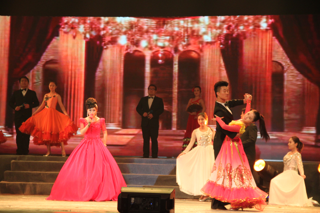 Eine Gruppe von Menschen steht auf einer Bühne mit Stufen und einem Hintergrundbild, beleuchtet von Scheinwerfern und mit einem Lautsprecher unten, was eine Tanzvorstellung suggeriert.