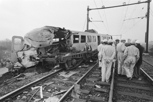 Eine Schwarz-Weiß-Szene eines Zugunglücks mit Menschen in Helmen drumherum, Strommasten, Bäumen, Gebäuden und einem klaren Himmel im Hintergrund.