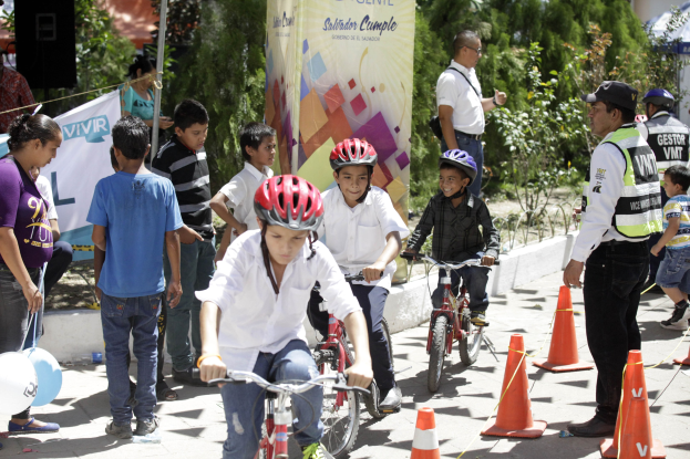 Kinder fahren Fahrräder auf einer Straße mit Verkehrshütchen, einige tragen Helme, andere stehen daneben, mit einem Banner, Bäumen und Gebäuden im Hintergrund.
