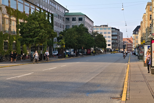 Eine Stadtstraße mit Fahrradfahrern, hohen Gebäuden, Bäumen, Laternen und Texttafeln unter einem sichtbaren Himmel.