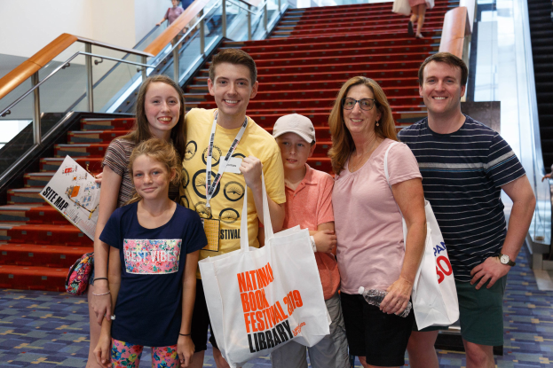 Eine Gruppe von Menschen, die vor einer Rolltreppe stehen und Taschen halten, mit Treppen, Geländern und einer Wand im Hintergrund.