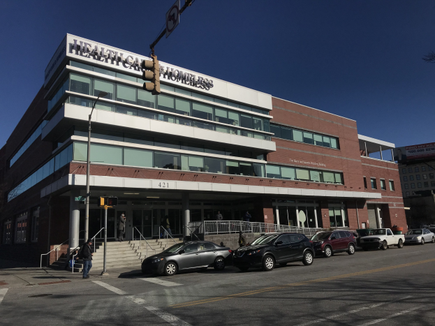 Großes Gebäude als New York City Health Care Homeless Center etikettiert mit parkenden Autos, Fußgängern, Straßenmöbeln und Bäumen unter einem klaren blauen Himmel.