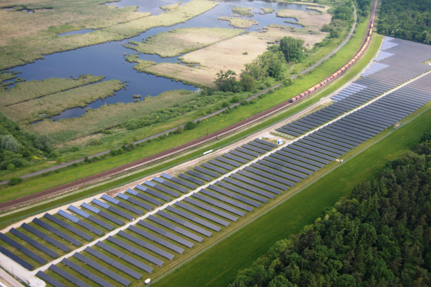 Luftaufnahme einer Solar-Farm mit Panelen, umgeben von Vegetation und Wasser, mit einem Zug auf einer nahen Schiene.