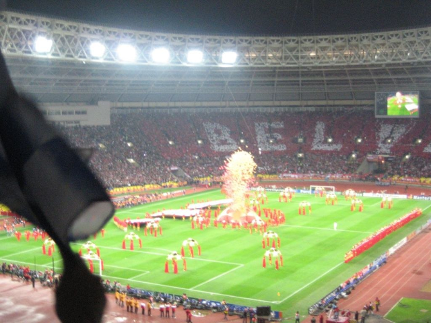 Ein Stadion mit Flutlicht, Eisengittern, Zuschauern, stehenden Personen am Boden, Werbetafeln, Anzeigetafeln, einer überwachungskamera, Fahnen und Deko.