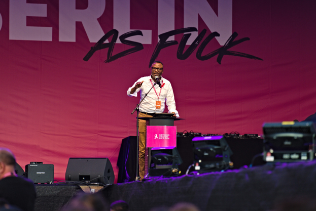Ein Mann in einem weißen Hemd und braunen Hosen steht auf einer Bühne und hält ein Mikrofon in der Hand, mit einem pinken Banner mit der Aufschrift "Berlin as Fuck" im Hintergrund und einer Menge davor.